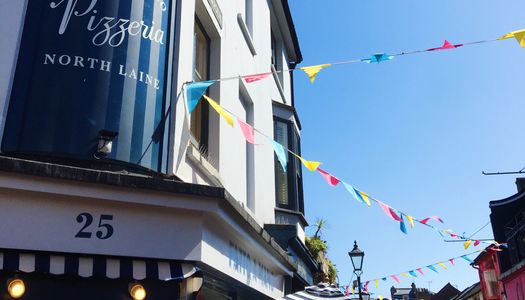 The exterior of Fatto A Mano on Gloucester Road next to Kensington Gardens in The North Laine. The picture shows the blue and white facade with bunting overhead against the back drop of the blue sky.