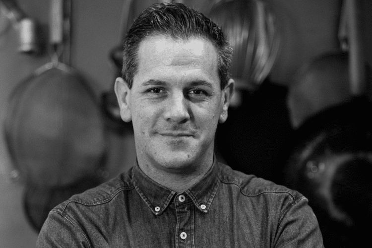 Oli Hyde from Flour Pot Bakery Brighton. Oli is folding his arms in a denim shirt. The picture is black and white. Oli is a handsome man. 