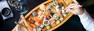 Sushi feast served on a wooden boat platter with people using chopsticks to pick pieces of sushi.