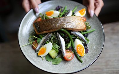 A fillet of fish sat on a bed of leafy salad with boiled eggs quartered. Served on a grey glazed ceramic plate.