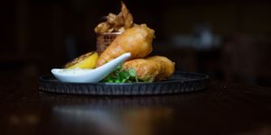 Battered fish pieces served with chips in a copper basket with a charred lemon half and microgreens.