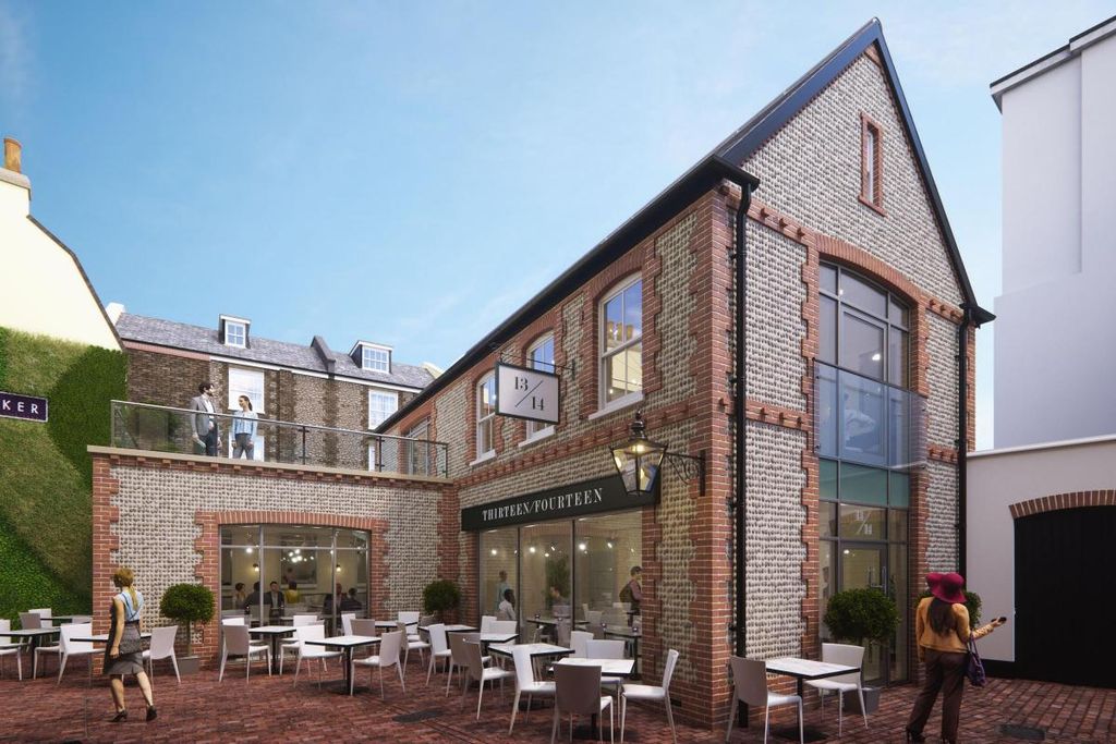 The Flint House Brighton. Pictured a brick facade, seating outside, an elevated upper terrace area and a person walking by in this area of the Brighton Lanes. It is a blue sky day and you can see a vertical plant wall to the left of the building.