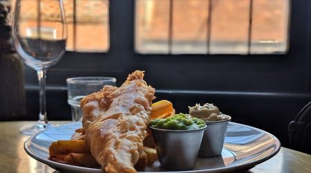 A large battered fish with chips, pots of mushy peas and tartare sauce served on a plate with a glass of wine.