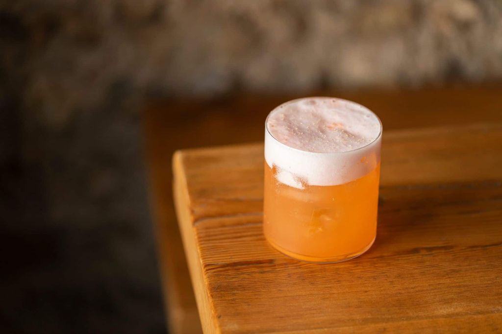 An orange coloured cocktail in a shot glass with a white foam head on a wooden table