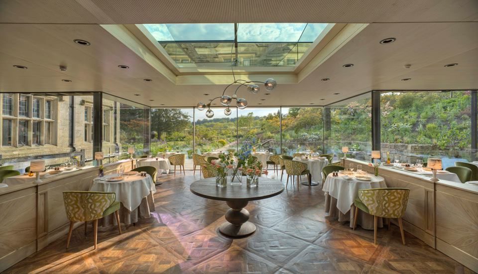 The dining room at Gravetye Manor with floor to ceiling walls all round showing a view of trees. Tables are dressed with white linen. There is an overhead window providing more light for this Sussex restaurant.