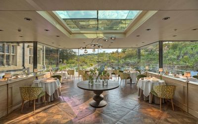 The dining room at Gravetye with floor to ceiling walls all round showing a view of trees. Tables are dressed with white linen. There is an overhead window providing more light for this Sussex restaurant.