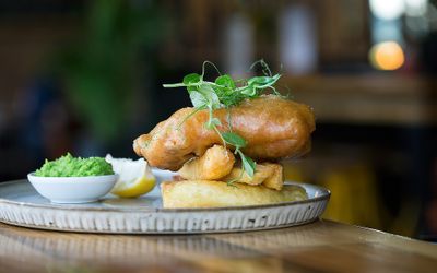 A piece of battered fish sat on a bed of chunky chips topped with pea sprouts and served with a small pot of mushy peas and a wedge of lemon.