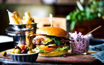 A colourful photo of a large burger stacked with burger, salad and cheese. Served with red cabbage slaw and a bucket of chips.
