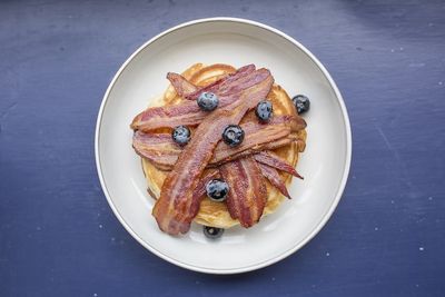 Pancakes with bacon and fresh blueberries served on a white plate sat on a purple table.