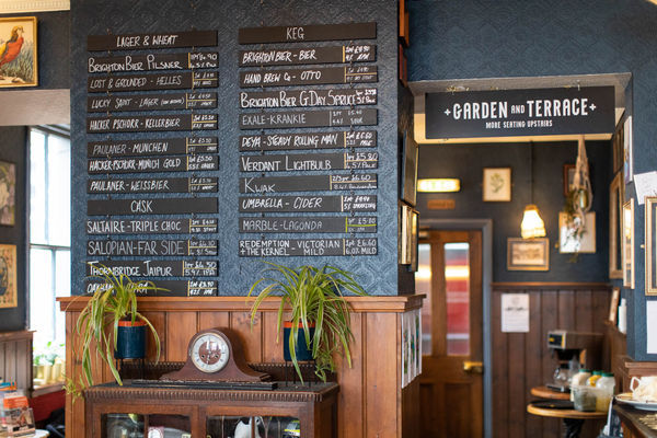 A dark green-blue wall is covered in blackboard strips each advertising a cask ale or beer. Wooden paneling covers the lower half of the wall and a vintage cabinet has two spider plants and a clock on top of it. To the left and right you can see through to the back of the bar and the garden