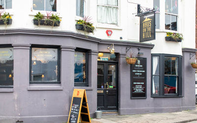 A pub front with a bay window and A frame sign to the left of the entrance and a swing sign to the right reading Haus on the Hill. The lower half of the building is painted a purple-grey with the window frames in black, the upper floor is white with window boxes full of foliage. Available for private dining Brighton