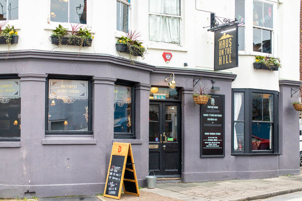 A pub front with a bay window and A frame sign to the left of the entrance and a swing sign to the right reading Haus on the Hill. The lower half of the building is painted a purple-grey with the window frames in black, the upper floor is white with window boxes full of foliage