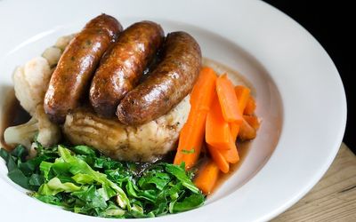 Sausages & mash with carrots, greens and gravy served in a white dish.