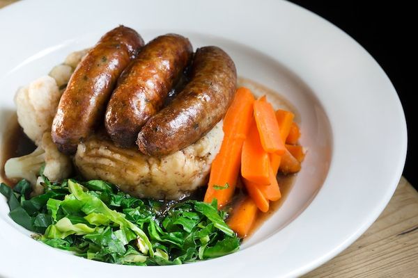 Sausages & mash with carrots, greens and gravy served in a white dish.