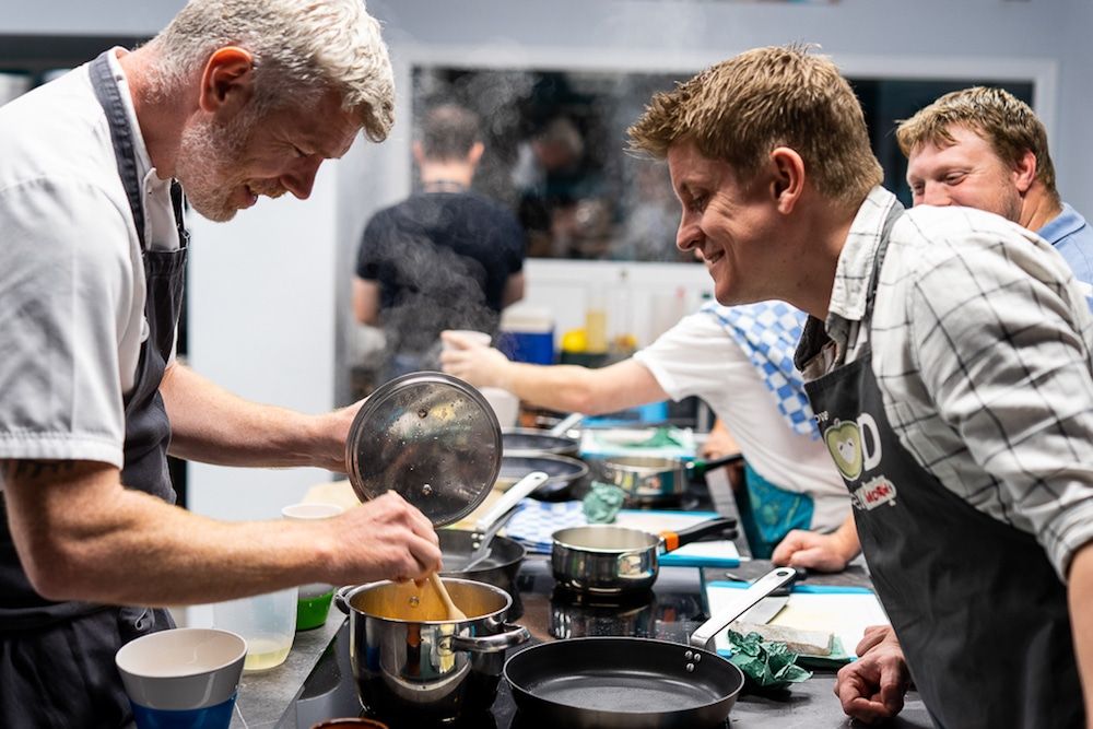 BHFP or Brighton Community Kitchen. Michael Bremner stirring a pot of food, with an onlooker looking over. 