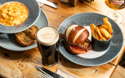 A flat-lay of a hearty pub lunch featuring a grilled chicken burger in a pretzel bun with chips, a bowl of mac and cheese, and a branded pint of Guinness from The Dorset.