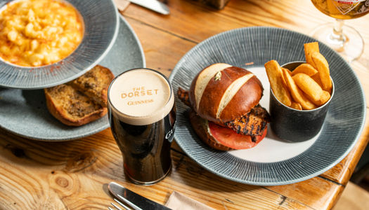A flat-lay of a hearty pub lunch featuring a grilled chicken burger in a pretzel bun with chips, a bowl of mac and cheese, and a branded pint of Guinness from The Dorset.