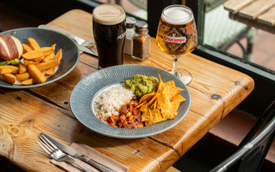 Close-up of loaded nachos topped with guacamole, sour cream, salsa, and jalapeños at The Dorset. In the background, a grilled chicken burger with chips and pints of Cruzcampo and Guinness sit on a wooden pub table.
