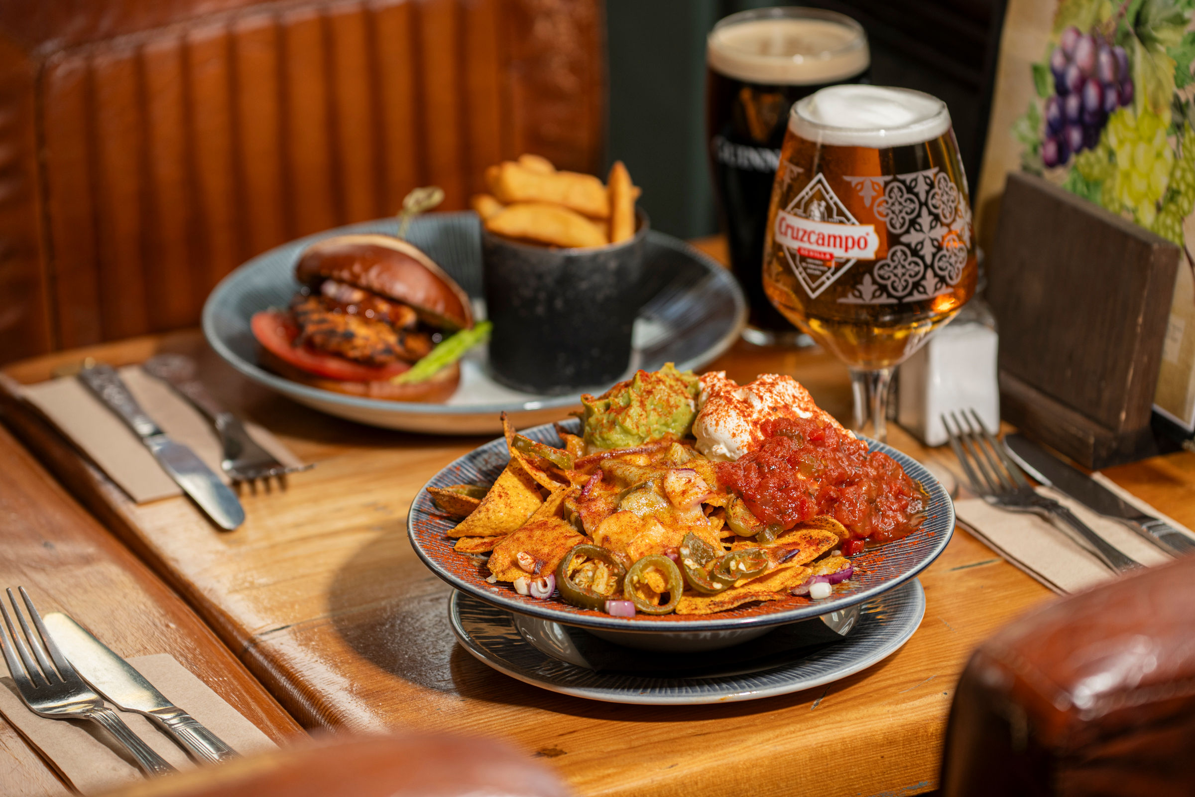 Close-up of loaded nachos topped with guacamole, sour cream, salsa, and jalapeños at The Dorset. In the background, a grilled chicken burger with chips and pints of Cruzcampo and Guinness sit on a wooden pub table.