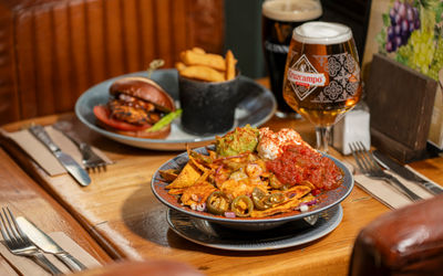 Close-up of loaded nachos topped with guacamole, sour cream, salsa, and jalapeños at The Dorset. In the background, a grilled chicken burger with chips and pints of Cruzcampo and Guinness sit on a wooden pub table.