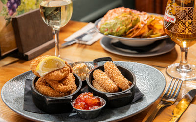 Fried prawns and mozzarella sticks served with sweet chilli dip and coleslaw, accompanied by a glass of white wine and Cruzcampo beer, set on a polished wooden table.
