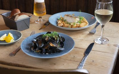 A bowl of moules marinière and a fish dish both served on a wooden table with a glass of wine and beer.