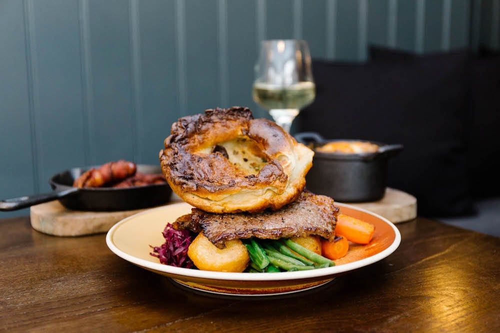 Huge Yorkshire pudding on top of a roast dinner at the Cleveland Arms Brighton. The food is on the wooden table with a dark blue backdrop. 