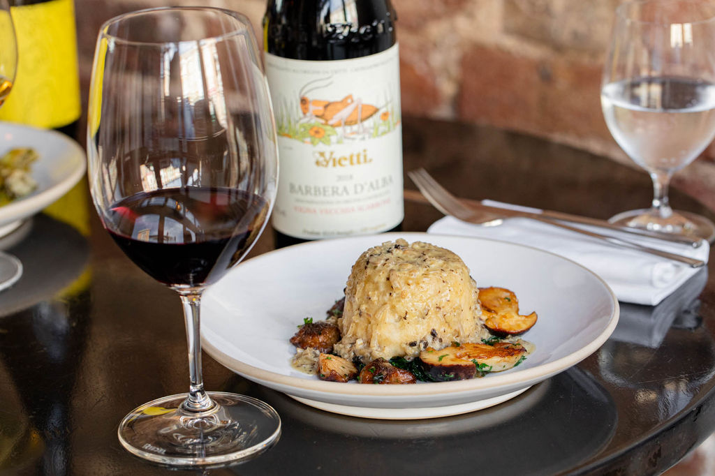 A white dish with food next to a glass of red wine and the bottle of wine in the background.