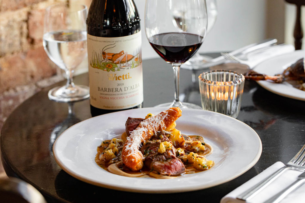 A white ceramic plate with a meat dish and a tea light in the background with a glass and bottle of red wine.