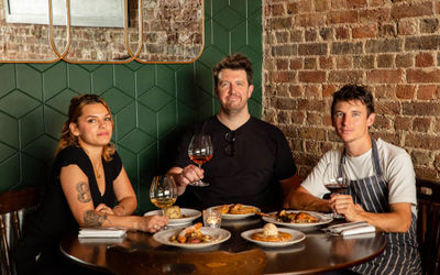 A team photograph of three people holding a glass of wine around a dinner table with brickwork showing on the walls.