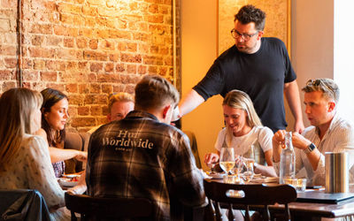 A group are seated at a table sharing food and wine while a man in a dark t-shirt pours wine, and exposed brick wall is behind them. Wild Flor is available for private dining in Brighton and Hove