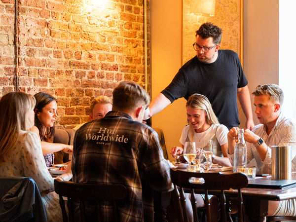 A group are seated at a table sharing food and wine while a man in a dark t-shirt pours wine, and exposed brick wall is behind them