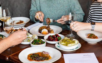 A close up of a table filled with food and wine glasses and around it the hands and torsos of diners all tucking in