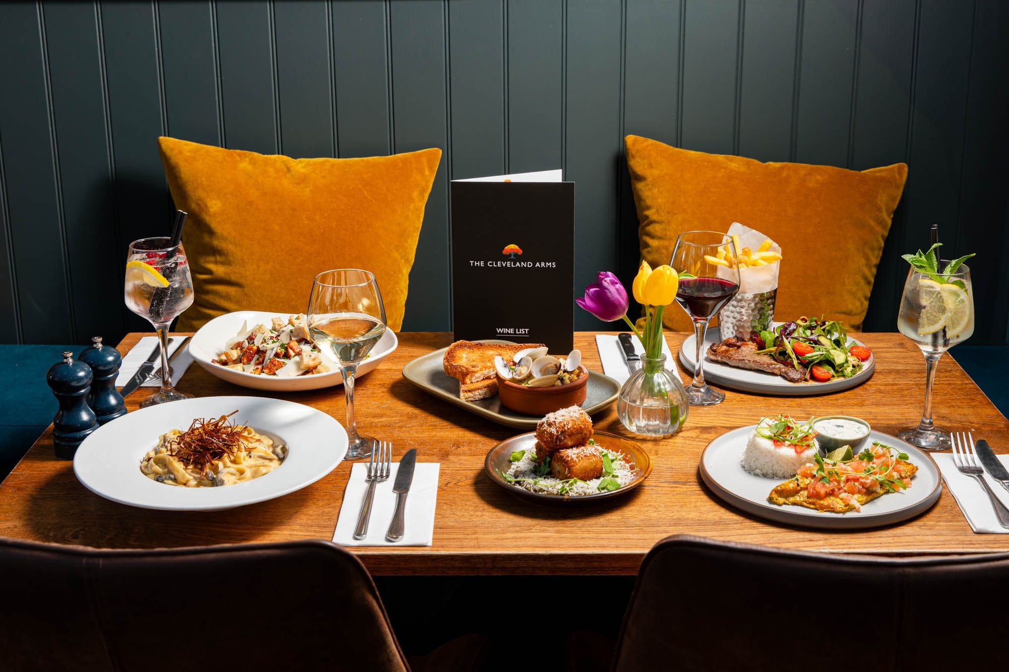 side shot of the brown wooden table laid out with the four main and two side dishes, served with glasses of wine at The Cleveland Arms in Brighton. 