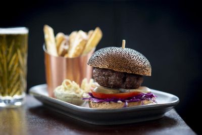 A rectangular plate serving a beef burger with shredded red cabbage inside a black sesame seed bun. Served alongside a metal pot of chunky fries and a pint of beer.