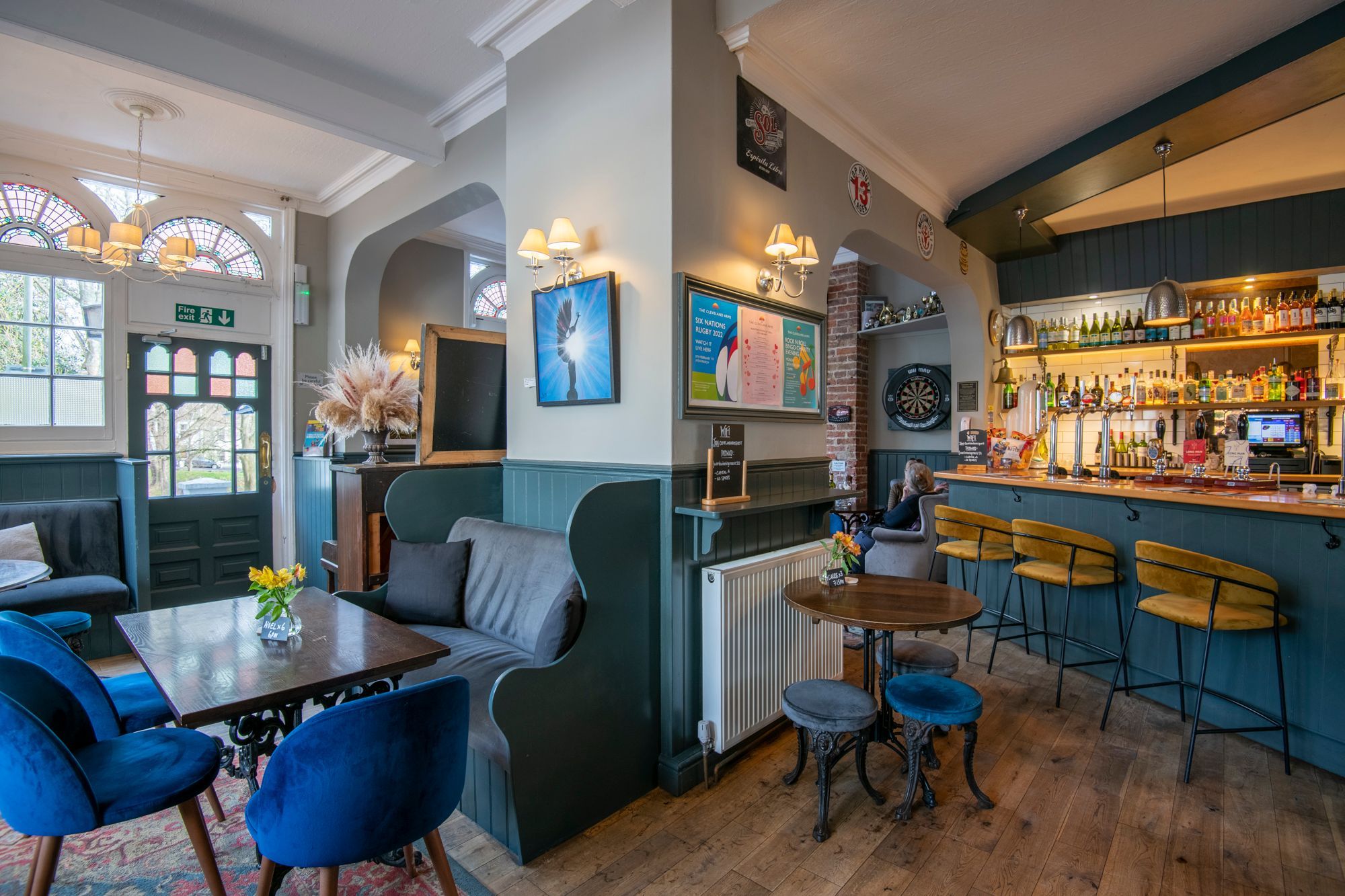 bar area and lounge area of Cleveland Arms Brighton. wooden tables with blue velvet seats, flowers on the table and a dartboard in the background. 