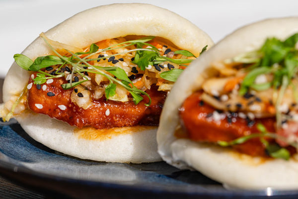 This close-up shot showcases a pair of beautifully crafted bao buns, filled with crispy, golden-brown protein coated in a rich, glossy sauce. The buns are topped with a fresh, crunchy slaw, a sprinkling of black and white sesame seeds, and delicate microgreens for added texture and flavour.