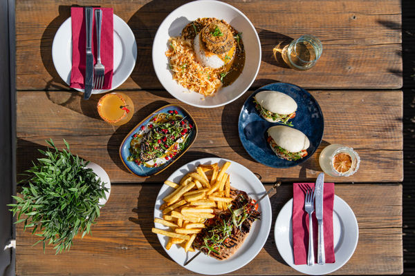 This overhead shot captures a beautifully arranged outdoor feast on a rustic wooden table, bathed in natural sunlight. The spread includes an array of vibrant and carefully plated dishes, from fluffy bao buns filled with crispy protein and fresh greens to a katsu-style curry dish with perfectly steamed rice and crunchy toppings. A plate of skewered grilled meat with golden fries sits alongside a colourful salad topped with pomegranate seeds. Complementing the meal are refreshing cocktails and glasses of wine, making for a picture-perfect al fresco dining experience.