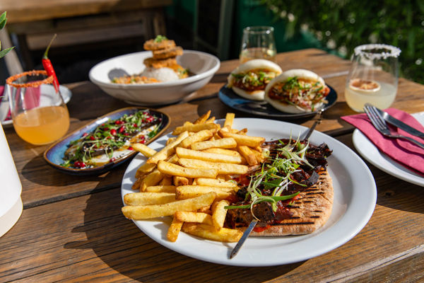 A beautifully plated feast featuring a grilled skewer served over warm flatbread, accompanied by golden crispy fries. In the background, a selection of vibrant dishes, including bao buns, fresh salads, and cocktails, add to the summery al fresco dining vibes.