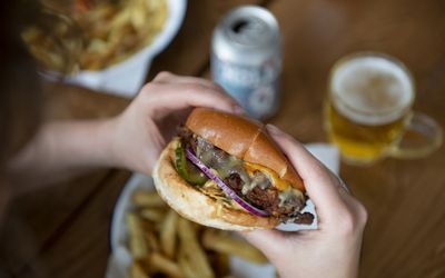 Hands holding a cheese burger with plates of fries and a can of craft beer in the background.