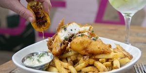 Pieces of battered fish on a bed pf fries with a squeeze of charred lemon and a little pot of tartare sauce.