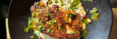 A close up photo of a cauliflower steak on a creamy sauce with fresh chopped herbs on a black dish.
