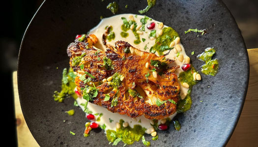 A close up photo of a cauliflower steak on a creamy sauce with fresh chopped herbs on a black dish.