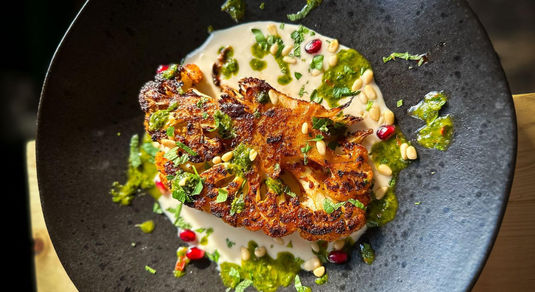 A close up photo of a cauliflower steak on a creamy sauce with fresh chopped herbs on a black dish.
