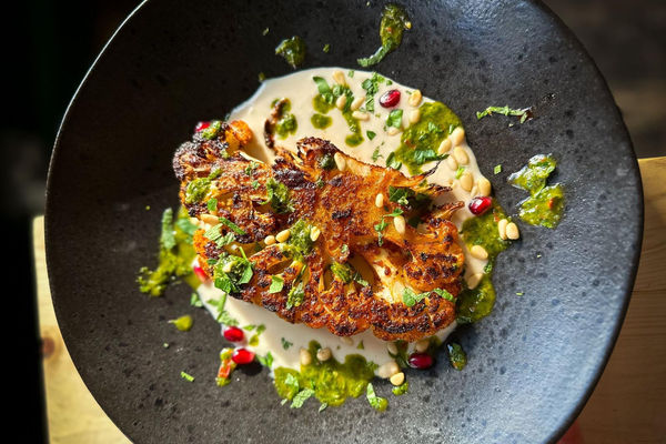 A close up photo of a cauliflower steak on a creamy sauce with fresh chopped herbs on a black dish.