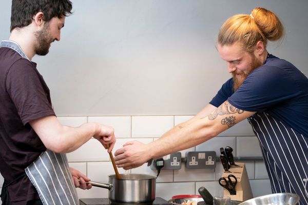 two men by the table cooking food, a man with the blue shirt and long ginger hair adding some spices to pot while man in the brown t shirt stirring the pot