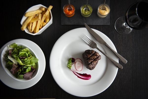 Steak dish with fries, sides and dips served on white dishes and a black table with a glass of red wine.
