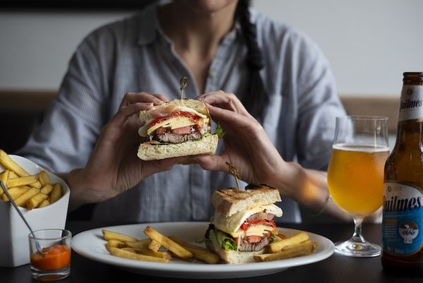 A person eating a beef burger that's been cut in half. Served on a white plate with fries and a glass of beer.