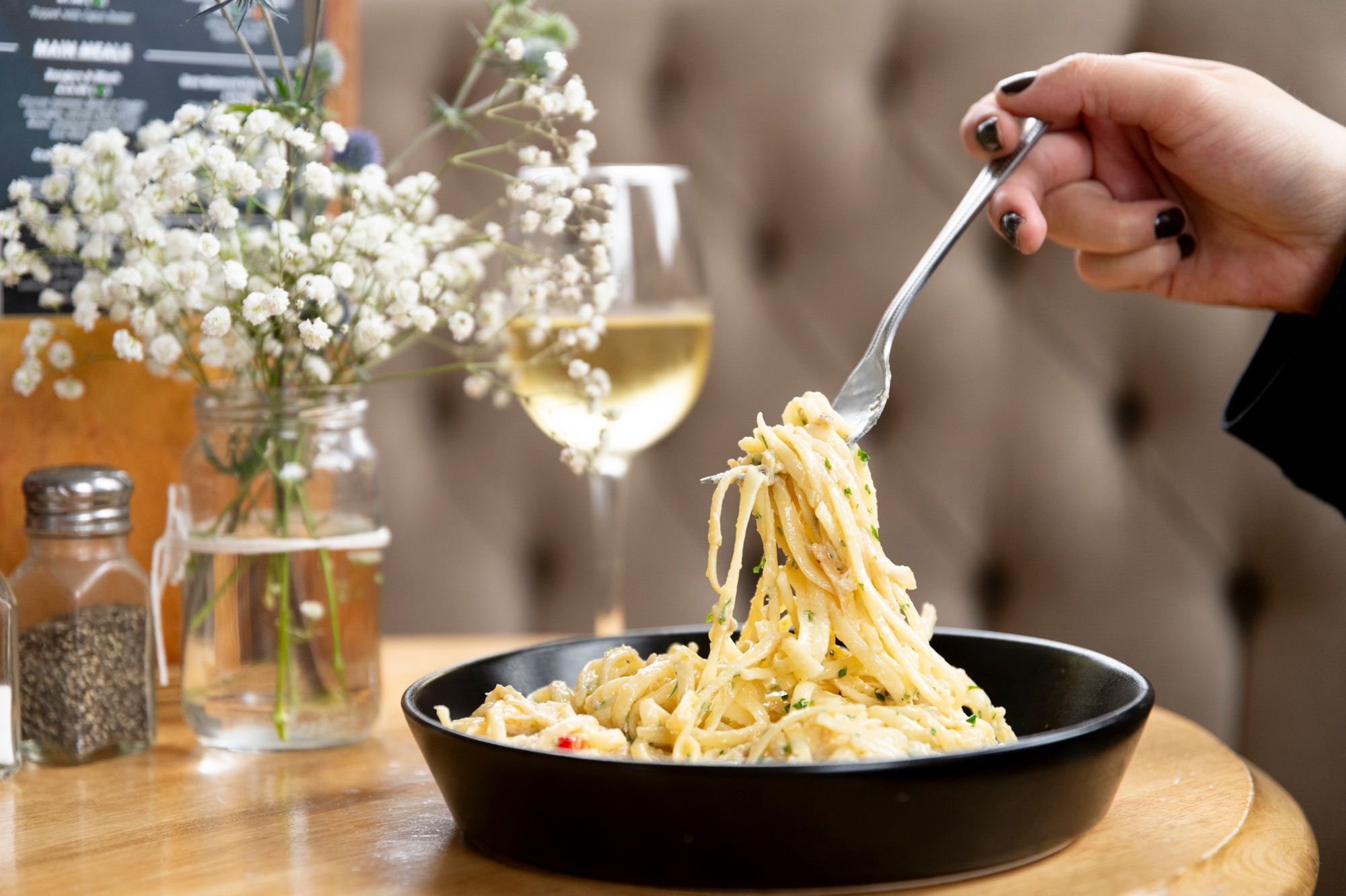 person taking pasta from the black plate with the fork. Food is served with the glass of white wine. The George Payne