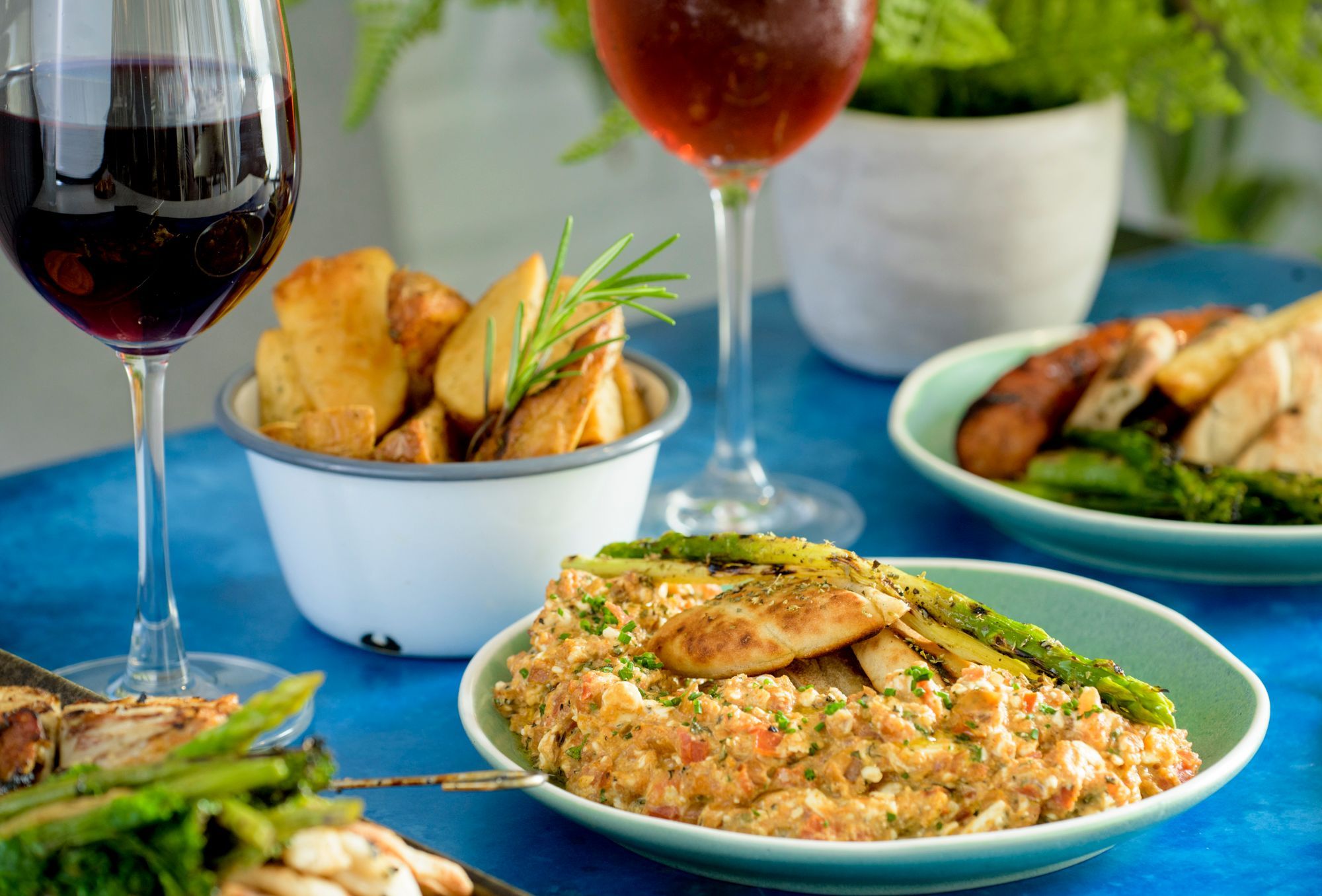 two glasses of red wine and three different dishes served on blue table - Nostos in Hove